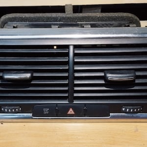 May include: A black car air vent with a silver trim. The vent has horizontal slats and two adjustable knobs on the bottom. The centre panel has an ESP OFF button and a hazard light button.