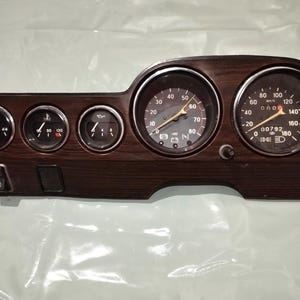 Könnte beinhalten: Ein Oldtimer-Armaturenbrett mit Holzmaserung. Es verfügt über mehrere Anzeigen, darunter einen Tachometer, eine Tankanzeige und eine Temperaturanzeige. Der Tachometer zeigt bis zu 160 mph an. Das Armaturenbrett hat auch einen Lichtschalter.