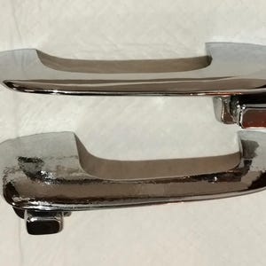 May include: Two chrome-plated door handles. The handles have a curved design with a rectangular base. The shiny metal reflects light, creating highlights and shadows. The handles are likely for a vehicle.