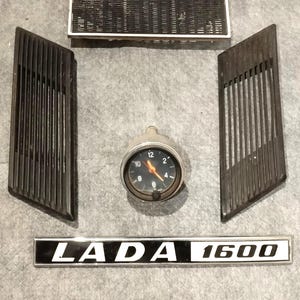 Puede incluir: Un emblema LADA 1600 en blanco y negro con un borde plateado. El emblema está sobre una superficie gris. También hay dos rejillas de metal negro y un reloj analógico redondo con agujas negras y una esfera blanca.