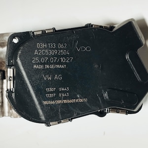 May include: A black plastic automotive throttle body component with a metallic silver base. The black surface features printed text including 03H 133 062, A2C53092504, 25.07.07/10:27, MADE IN GERMANY, VW AG, 13307 01443, 13307 0443, and 015866/205/150607/00050.