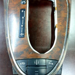 Puede incluir: Un panel de cambio de marchas de coche de madera marrón con un borde plateado. El panel tiene una palanca de cambios, un botón C/S y tres botones con símbolos para el freno de estacionamiento, el control electrónico de estabilidad y la asistencia de estacionamiento.