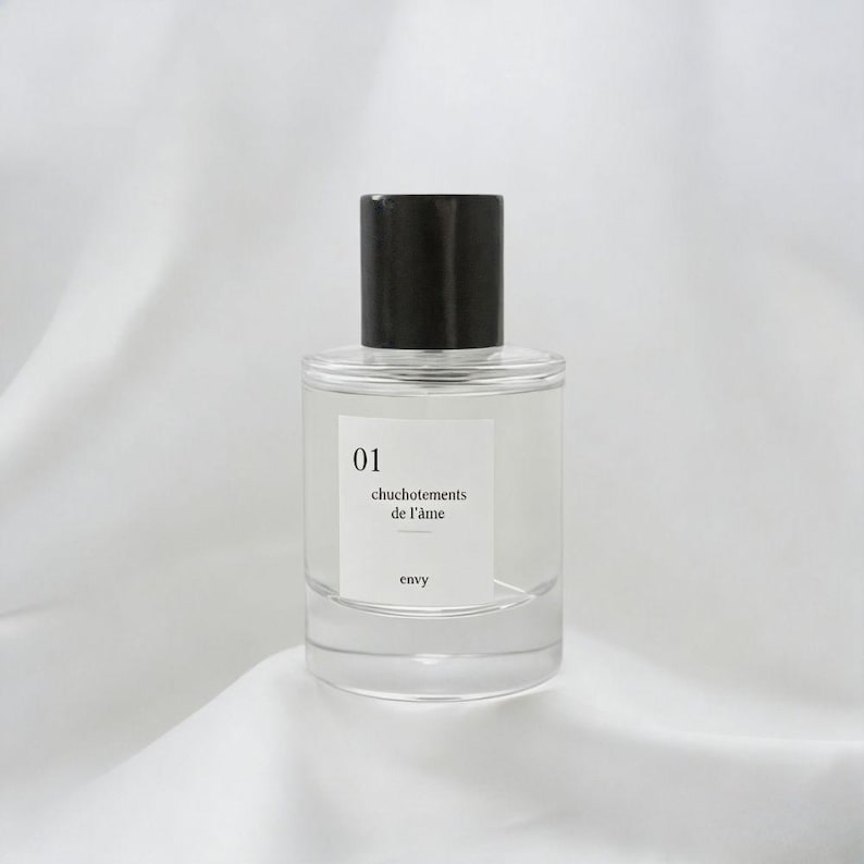 Pu&ograve; includere: Un flacone di profumo trasparente con tappo nero. Il flacone ha un'etichetta bianca con il numero "01", il testo "chuchotements de l'&acirc;me" e la parola "envy". &Egrave; posto su uno sfondo bianco drappeggiato.