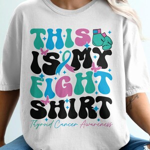 May include: White t-shirt with the text "THIS IS MY FIGHT SHIRT" in a retro font, with teal, pink, and black colors. The shirt also features a teal ribbon, butterflies, and boxing gloves. The phrase "Thyroid Cancer Awareness" is printed below.