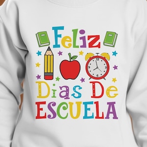 Puede incluir: Sudadera blanca con el texto "Feliz Dias De Escuela" en letras de colores. El texto está rodeado de estrellas de colores, una manzana roja, un despertador rojo, un lápiz amarillo y dos libros cerrados.