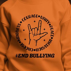 May include: Orange sweatshirt with a black graphic of a hand making the American Sign Language symbol for 'I love you'. The graphic is surrounded by a circle with the words 'Choose Kindness, Courage, Unity, Peace, Inclusion'. The text '#End Bullying' is below the graphic.