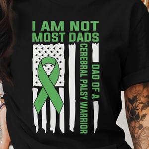 Op de afbeelding: Zwart T-shirt met een groen en witte afbeelding. De afbeelding toont een groen lint, een gestileerde Amerikaanse vlag en de tekst "I AM NOT MOST DADS" en "DAD OF A CEREBRAL PALSY WARRIOR".
