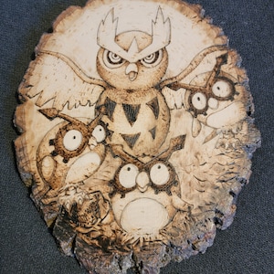 May include: A woodburned image of a large owl with three smaller owls surrounding it. The owls are all facing forward with their eyes wide open. The wood is a light brown color and the owls are burned into the wood in a dark brown color.