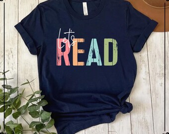 May include: A navy blue short-sleeve t-shirt featuring the text "let's READ" printed in a distressed, multi-colored font. The word "let's" is in white script, while the letters in "READ" are colored in pink, orange, light blue, and green.