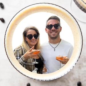 Puede incluir: Dos personas sonriendo, sosteniendo cócteles naranjas en copas con tallo. Ambos llevan gafas de sol con forma de corazón. Las gafas de sol de la mujer tienen la palabra "FAVOURITE". Las gafas de sol del hombre tienen la palabra "BEN". La imagen está enmarcada por un diseño circular.