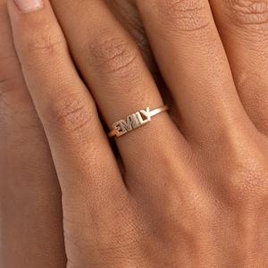 Puede incluir: Un anillo dorado con el nombre "EMILY" en letras mayúsculas. El anillo es delgado y delicado, con las letras recortadas del metal. Se lleva en un dedo, mostrando un diseño minimalista.