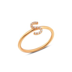 Puede incluir: Un anillo dorado con la letra "S" como elemento principal del dise&ntilde;o. La letra est&aacute; incrustada con peque&ntilde;as piedras preciosas transparentes. El anillo tiene una banda simple y elegante.