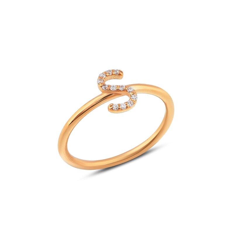 Puede incluir: Un anillo dorado con la letra "S" como elemento principal del dise&ntilde;o. La letra est&aacute; incrustada con peque&ntilde;as piedras preciosas transparentes. El anillo tiene una banda simple y elegante.