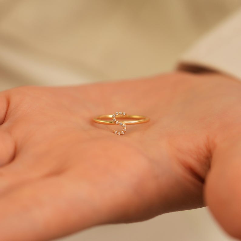 Puede incluir: Un anillo dorado con la letra "S" incrustada con peque&ntilde;as piedras preciosas transparentes. El anillo es delicado y se encuentra en la palma de una persona, mostrando sus finos detalles. Es una pieza de joyer&iacute;a.