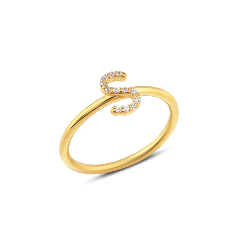 May include: A gold-colored ring featuring the letter "S" encrusted with small, clear gemstones. The ring has a delicate band and a stylized design, perfect for adding a personalized touch to any outfit.