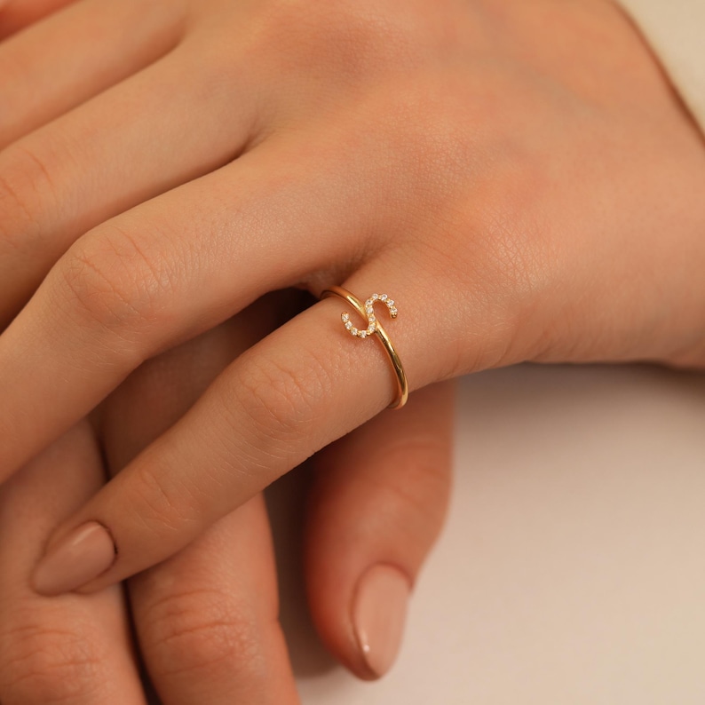 Puede incluir: Un anillo dorado con la letra "S" adornada con peque&ntilde;as piedras preciosas transparentes. El anillo es delicado y se asienta en un dedo, destacando su dise&ntilde;o minimalista y sus detalles brillantes. La banda del anillo es fina y pulida.
