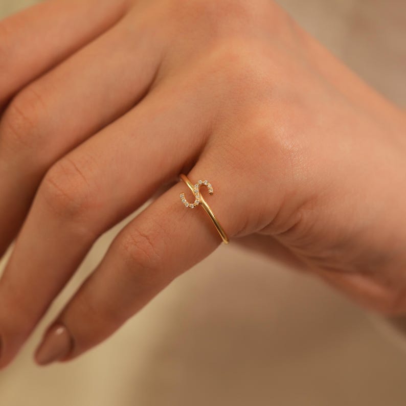 Puede incluir: Un delicado anillo de oro con la letra "S" incrustada con peque&ntilde;as y brillantes gemas. El anillo es fino y elegante, dise&ntilde;ado para ser usado en un dedo. El anillo se muestra en una mano con esmalte de u&ntilde;as neutro.