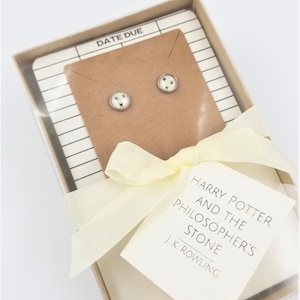 May include: A white gift box with a yellow ribbon and a card that reads "Harry Potter and the Philosopher's Stone J.K. Rowling". The box contains a pair of silver stud earrings with a white star design.
