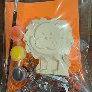 May include: A craft kit containing a white plaster lion figurine, a black-handled paintbrush, and three small circular paint pots in orange, yellow, and black. The items are arranged on an orange card inside a clear plastic bag with silver tinsel.
