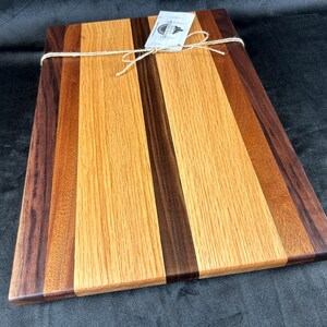 May include: A rectangular wooden chopping board with a mix of wood shades, such as dark brown, medium brown, and light brown. The board is tied with twine and has a small tag attached. This handcrafted board is ideal for kitchen use.