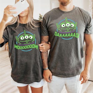 May include: A couple wearing matching gray t-shirts with a green alien graphic. The woman's shirt says "MOOOOODOOOM!" and the man's shirt says "DAAAAAAAAAD!"