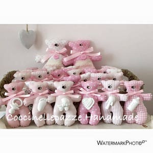 Puede incluir: Colecci&oacute;n de peluches de ositos de peluche rosas y blancos con lazos rosas. Algunos ositos tienen lunares o estampados de cuadros. Varios ositos sostienen accesorios para beb&eacute;s. El texto "Coccinellepazze Handmade" es visible.