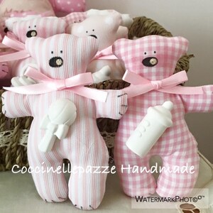 Puede incluir: Osos de peluche a rayas y cuadros rosas y blancos con lazos de cinta rosa. Un oso sostiene un sonajero blanco, el otro un biber&oacute;n blanco. El texto "Coccinellebazze Handmade" es visible.