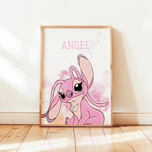 May include: A framed print showing a pink cartoon character with large ears and big eyes. The word "ANGEL" is written in pink above the character. The background has pink and purple watercolour-like accents.