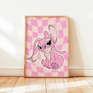 May include: A framed art print featuring a pink cartoon character with large eyes and long ears, set against a pink and white checkered background. The artwork is in a wooden frame and is displayed on a wooden floor.