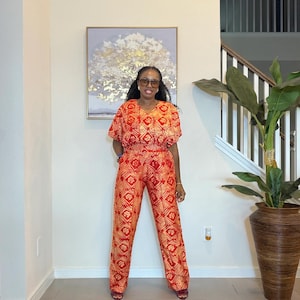 May include: A two-piece outfit consisting of a short-sleeved V-neck top and matching high-waisted trousers, both featuring a vibrant orange and cream geometric pattern with repeating diamond shapes and small floral motifs throughout the fabric.