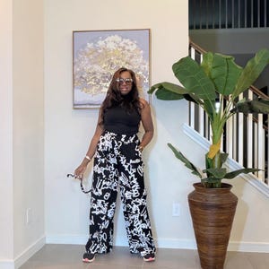 May include: Black and white palazzo pants with a bold, abstract print of white lettering. The pants are paired with a black sleeveless top. The outfit is completed with sunglasses and sneakers. A large decorative plant in a brown vase is in the background.