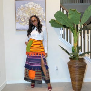 Könnte beinhalten: Ein langer, farbenfroher Rock mit hohem Schlitz, der horizontale Streifen in Orange, Gelb und Blau sowie Fransen an den Kanten aufweist. Der Rock wird mit einem weißen Langarmhemd kombiniert. Der Rock hat ein Patchwork-Design mit grünen und orangen Akzenten.