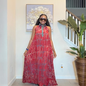 May include: A red halter-neck maxi dress with a paisley pattern. The dress features a high neckline with a decorative trim and a flowing, floor-length skirt. The dress is worn with sunglasses and a bracelet.