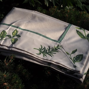 May include: A folded off-white tablecloth featuring a subtle tonal damask pattern, accented with a thin sage green border and a printed botanical design of green rosemary sprigs and leafy stems across the fabric surface.