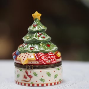 Puede incluir: Una caja de baratijas con temática navideña con forma de árbol. El árbol verde tiene detalles rojos y blancos y una estrella amarilla en la parte superior. La base es una caja redonda con un patrón festivo de hojas de acebo, corazones rojos y lazos. La caja está adornada con rayas rojas y blancas.