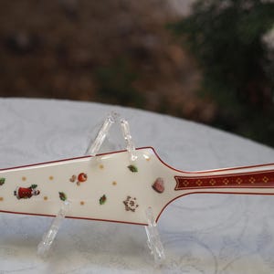 Può includere: Un servizio per torta in ceramica bianca con un bordo rosso e dorato. Il servizio è decorato con un motivo natalizio festoso con un omino di pan di zenzero, un cuore e agrifoglio.