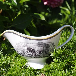 Puede incluir: Una salsera de porcelana blanca con una escena pastoral en blanco y negro de vacas pastando en un campo. El borde está decorado con una fina banda dorada y un patrón geométrico griego negro. Tiene un asa curva y una base de pedestal.