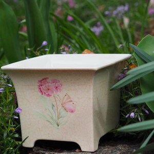 May include: A square, off-white ceramic planter with a floral and butterfly design. The planter has a slightly flared shape and sits on four small feet. The design features pink flowers and a butterfly. The planter is surrounded by greenery.