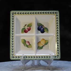 May include: A white ceramic serving dish with four compartments. Each compartment features a different fruit design: cherries, plums, a rose, and lemons. The dish has a green and white border with a repeating arrow pattern.