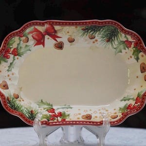 Può includere: Un piatto da portata ovale in ceramica con bordo smerlato e rifinitura rossa. Il bordo presenta un motivo festivo di rami di pino verdi, bacche di agrifoglio rosse, un fiocco rosso e piccoli biscotti marroni a forma di cuore su fondo crema.