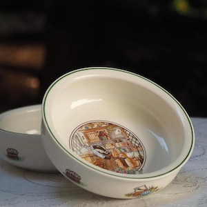 Puede incluir: Dos cuencos de cerámica blanca con ribete verde. El cuenco más grande tiene una ilustración colorida del interior de una panadería con personas dentro. El cuenco más pequeño es liso.