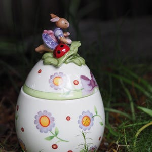 Op de afbeelding: Een witte, eivormige keramische container met een bloemenpatroon en een deksel met een klein konijnenbeeldje dat op een lieveheersbeestje rijdt.