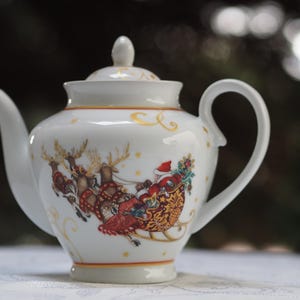 Pode incluir: Bule de porcelana branca com uma cena de Natal representando o Papai Noel em um trenó puxado por renas. A chaleira tem uma borda dourada e uma tampa.