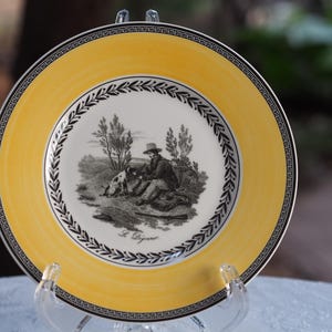 Puede incluir: Un plato redondo de cerámica con un borde amarillo brillante que presenta un diseño de greca negra. El centro es blanco con una ilustración en blanco y negro de una persona y un perro en un campo, con el texto Le Déjeuner impreso debajo.