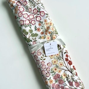 May include: A white fabric table runner featuring an embroidered floral pattern in shades of pink, yellow, and green. The runner is rolled and tied with a white ribbon, including a tag labeled "FLOWERVINE RUNNER" with dimensions 18 x 108 inches.