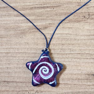 May include: A star-shaped pendant necklace. The pendant is a deep purple with white spiral and edge detailing, and a dark blue border. The necklace cord is a matching dark blue. The pendant is approximately 1.5 inches wide.