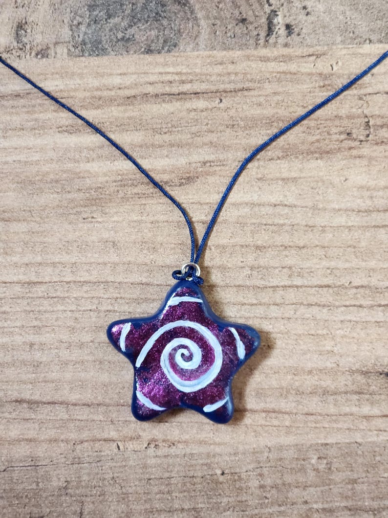 May include: A star-shaped pendant necklace. The pendant is a deep purple with white spiral and edge detailing, and a dark blue border. The necklace cord is a matching dark blue. The pendant is approximately 1.5 inches wide.