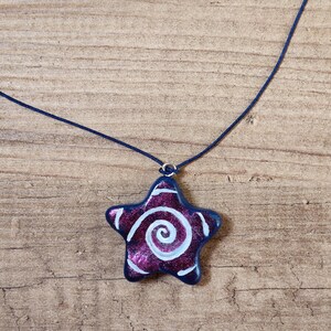May include: A star-shaped pendant necklace. The pendant is a deep purple with a white spiral design and a dark blue border. The necklace cord is a matching dark blue.