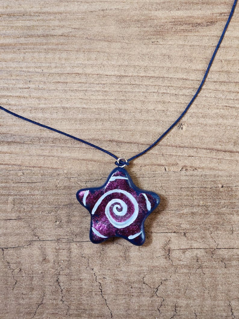 May include: A star-shaped pendant necklace. The pendant is a deep purple with a white spiral design and a dark blue border. The necklace cord is a matching dark blue.