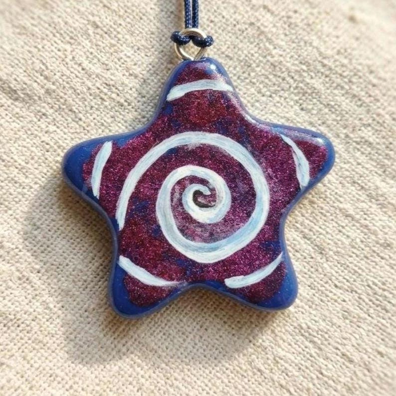 May include: A star-shaped pendant with a blue border and a swirling white design on a glittery purple background. The pendant is suspended from a dark blue cord, creating a unique and eye-catching accessory.